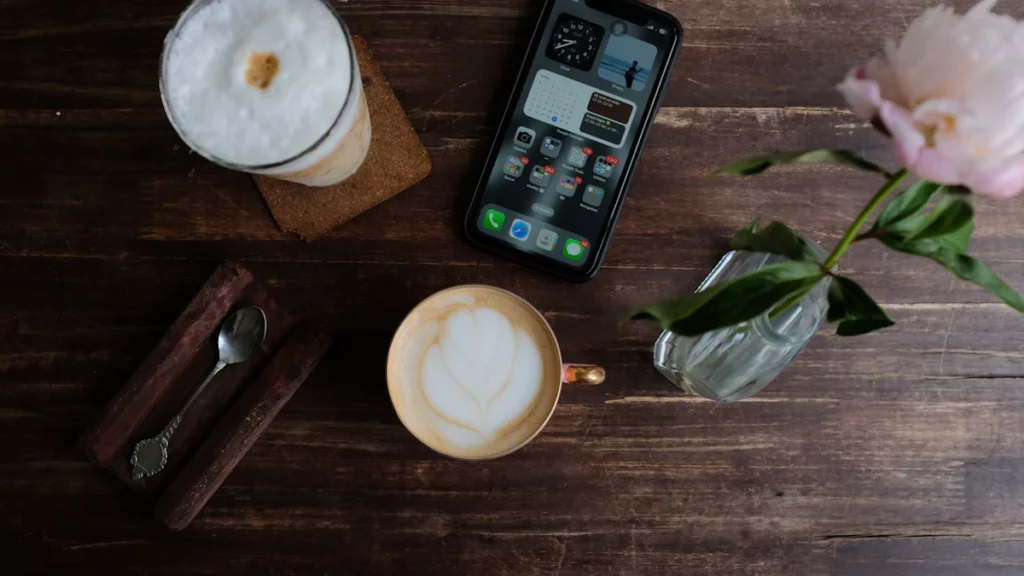 木のテーブルに置かれたカフェラテとスマートフォン、ピンクの花
